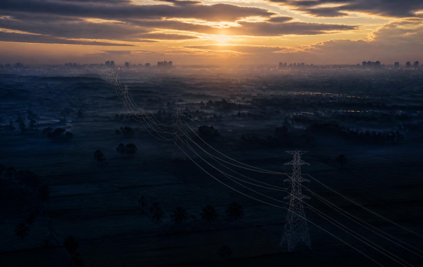 Transmission lines crossing a plain towards a distant city skyline at dawn